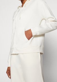 Felpa bianca con zip, cappuccio, maniche lunghe, tasche frontali e polsini a costine. Texture morbida e vestibilità comoda, abbinata a pantaloni coordinati.