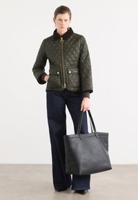 Gequilted olijfgroene jas met een bruine kraag, gouden rits en voorzakken; gecombineerd met donkere wijde jeans en een zwarte handtas.