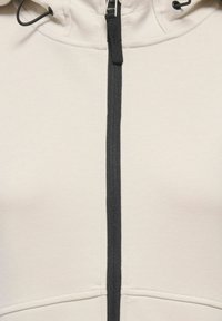 Veste beige clair avec une fermeture éclair noire centrée et une languette noire, avec des cordons près du bord supérieur du col.