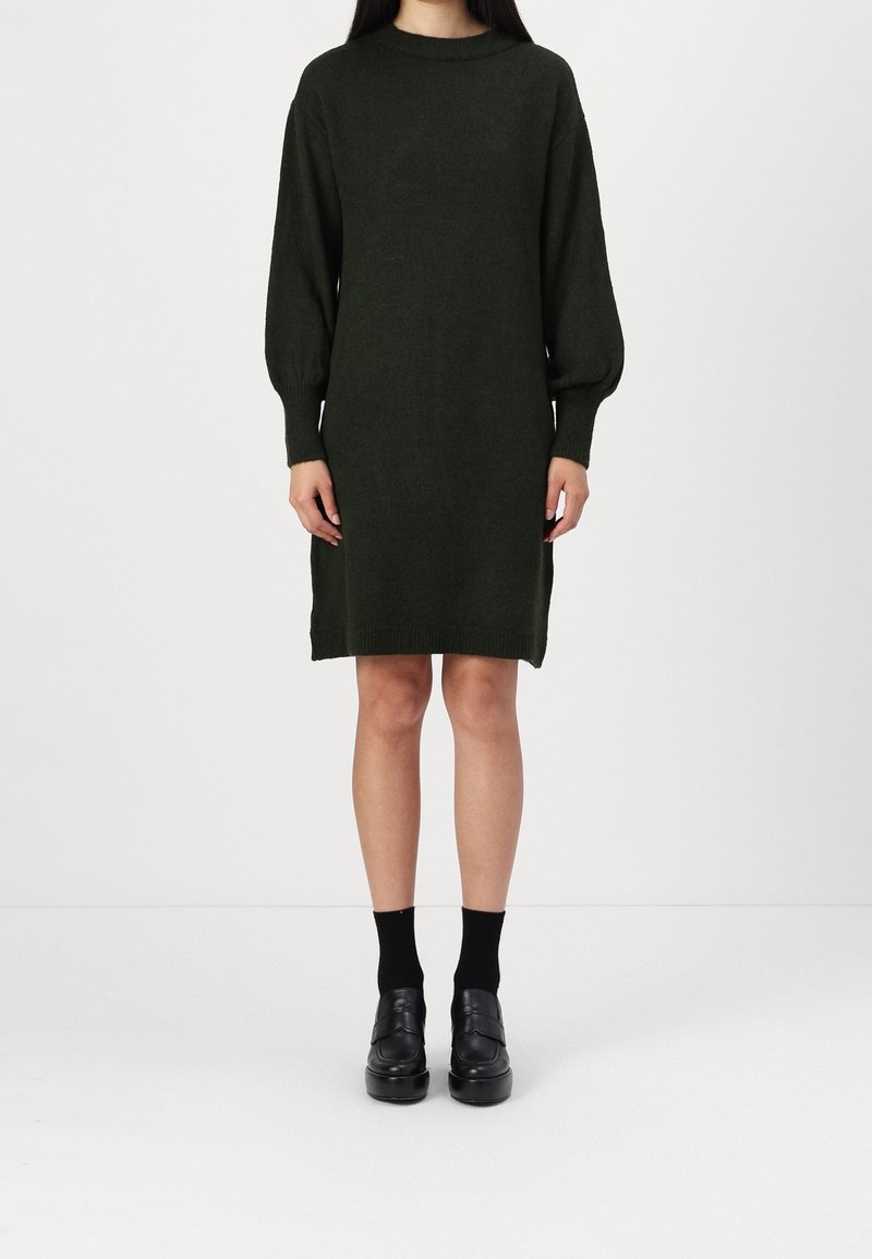 Femme portant une robe pull en maille vert foncé à manches longues, avec des chaussettes noires et des mocassins à plateforme noirs, debout sur un fond blanc.