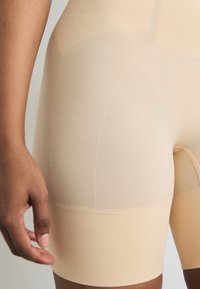 Sima textúrájú, bézs színű formáló rövidnadrág, bordázott derékkal, közép-thigh hosszúsággal és varrás nélküli kialakítással a kényelem érdekében.