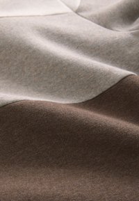 Teksturirana tkanina z gladkim prehodom svetlo sive, bež in temno rjave barve, z vidnimi detajli šivov in mehko padajočo tkanino.