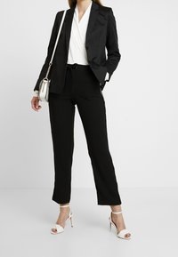 Blazer negro ajustado combinado con top blanco envolvente y pantalones negros. Sandalias de tacón alto blancas puntiagudas y un pequeño bolso cruzado.
