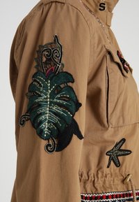 Brązowa kurtka z zielonymi i czarnymi haftowanymi liśćmi na rękawie, z dodatkowymi ozdobnymi detalami i ściągnięciem w talii.