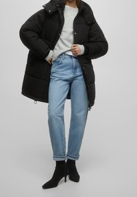 Manteau matelassé noir avec un col montant, pull gris, jean en denim bleu clair, et bottines noires à talons hauts. Textures lisses, coupe décontractée.