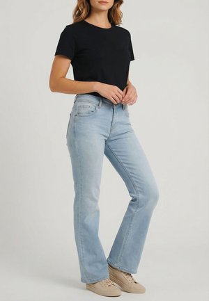 Donna in piedi che indossa una maglietta nera a maniche corte, jeans bootcut azzurri e sneakers beige su uno sfondo neutro.