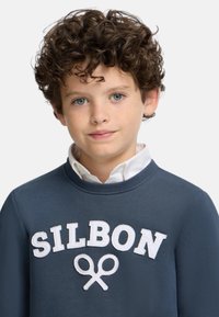 Sudadera azul marino con "SILBON" en letras bordadas blancas y un gráfico de tijeras. Llevada sobre una camisa blanca con cuello. Presenta un cuello redondo.