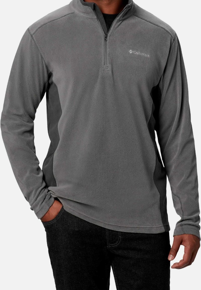 Homme portant un pull polaire gris à manches longues avec une fermeture éclair à l’avant et un pantalon noir, main dans la poche.