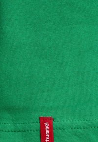 Grøn bomuldsstof med en glat tekstur. Har et rødt mærke med brandnavnet nederst på kanten. Enkel og udekoreret design.