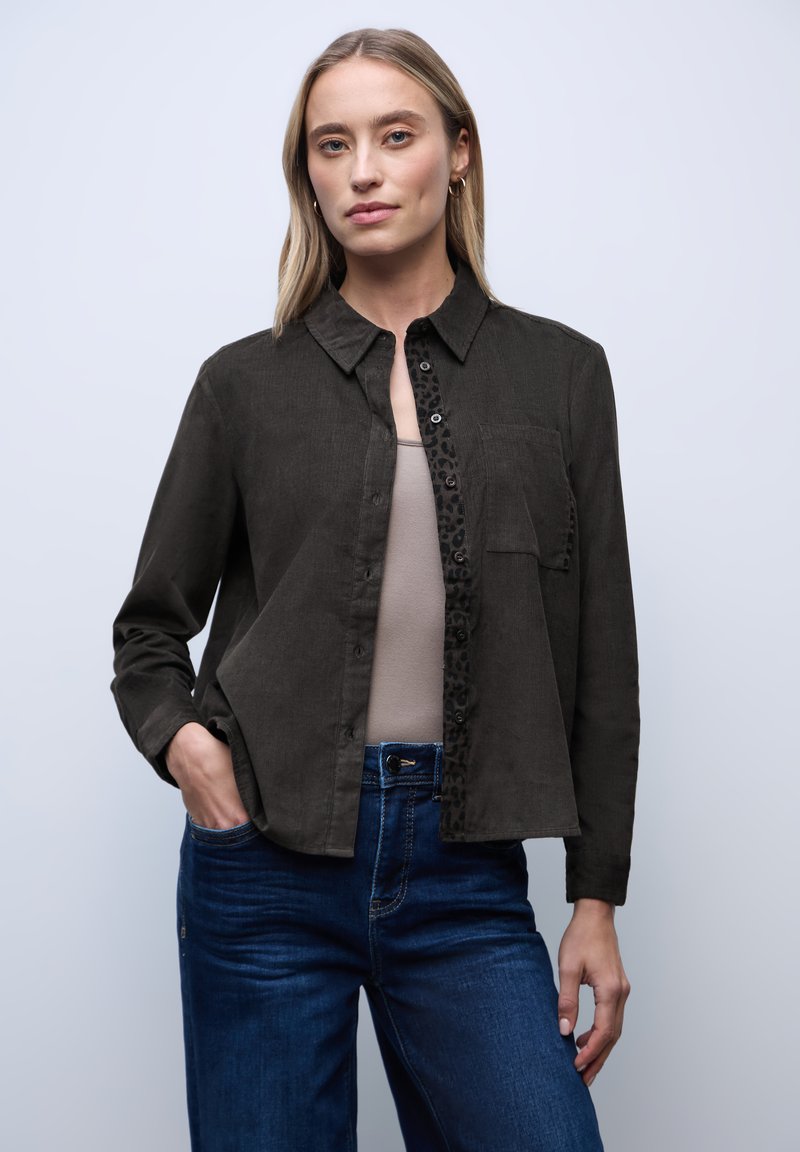 Vrouw met blond haar, die gouden oorbellen draagt, een donker knoopshirt over een beige top, blauwe spijkerbroek, staand met één hand in de zak.