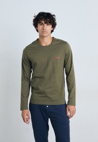 T-shirt à manches longues vert olive avec un col rond et le logo rouge "HUGO" sur la poitrine, associé à un pantalon bleu foncé. Texture lisse.