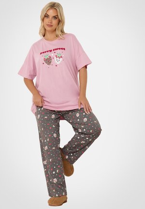 Kvinde iført pink "Berry sweet" t-shirt, grå pyjamasbukser med jordbærprint og brune hjemmesko poserer foran en helt hvid baggrund.