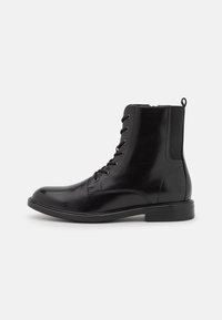 Botas de tobillo de cuero negro con cordones, punta redondeada, suelas resistentes y paneles laterales elásticos. Presentan ojales de metal y un lazo trasero para facilitar el calzado.