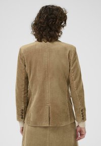 Blazer en velours côtelé marron avec une coupe ajustée, des coutures détaillées, une fente centrale et des boutons décoratifs sur les manches. Tissu texturé visible.