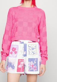 Ljusrosa stickad tröja med rutigt mönster, kombinerad med vita shorts med flerfärgade blommönster i olika rutor.