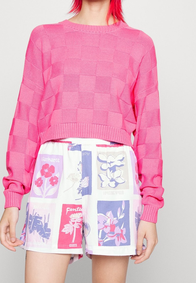 Ljusrosa stickad tröja med rutigt mönster, kombinerad med vita shorts med flerfärgade blommönster i olika rutor.