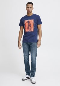 Mörkblå bomullströja med en orange grafik av palmer, omgiven av text. Bärs med medelblå jeans och grå sneakers.