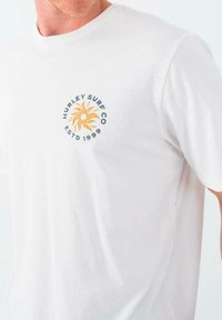 Camiseta blanca de algodón de manga corta, con un logo circular azul y amarillo que presenta un diseño de sol y el texto "Hurley Surf Co ESTD 1999."