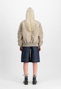 Beige bomber jacket with ribbed cuffs, paired with dark denim shorts. Black lace-up boots complete the outfit. Long light-colored hair visible.