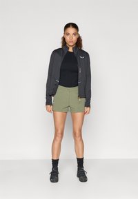 Μαύρο αθλητικό μπουφάν με ψηλό γιακά, συνδυασμένο με ελαιοπράσινα σορτς. Το μπουφάν διαθέτει φερμουάρ και αντίθετες ραφές. Υφασμα με υφή.