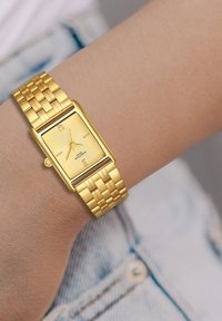 Zelta taisnstūra pulkstenis ar teksturētu ciparnīcu un minim�ālistiskiem marķieriem, aprīkots ar austu metāla siksniņu. Pulkstenis ir uz rokas.