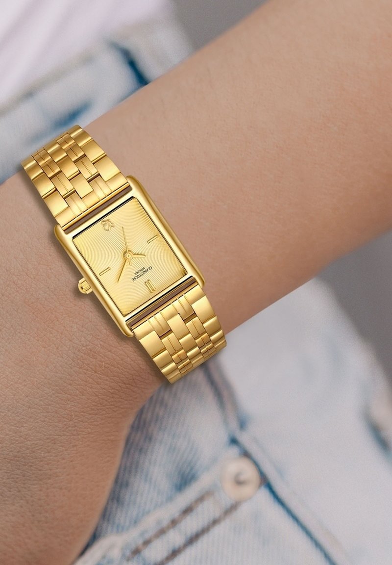 Zelta taisnstūra pulkstenis ar teksturētu ciparnīcu un minimālistiskiem marķieriem, aprīkots ar austu metāla siksniņu. Pulkstenis ir uz rokas.