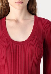 Haut en maille côtelée rouge avec un décolleté rond et un tissu texturé. Le design présente une silhouette ajustée avec des stries verticales.