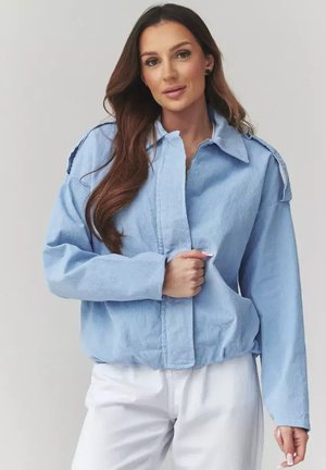 Kobieta o długich brązowych włosach, ubrana w jasnoniebieską kurtkę na zamek i białe spodnie, trzymająca kurtkę przy zamku, neutralne tło.