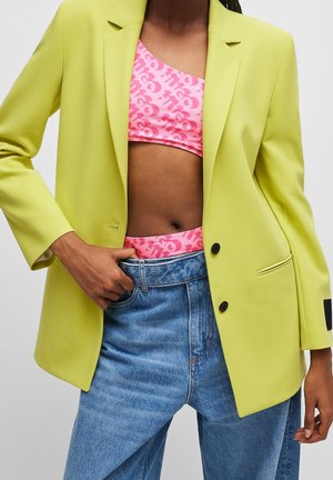 Person trägt einen limonengrünen Blazer über einem rosa gemusterten Crop-Top und hoch taillierten blauen Jeans, mit einer Hand in der Tasche.