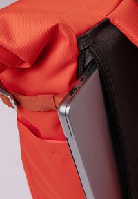 Orange tyg ryggsäck med rulltopp och svart dragkedja, som rymmer en silver laptop, visar texturerat material och justerbar rem.