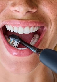 Brosse à dents électrique avec un manche noir et des poils blancs, positionnée près des dents. Bouche grande ouverte montrant des dents blanches et saines ainsi que des gencives roses.