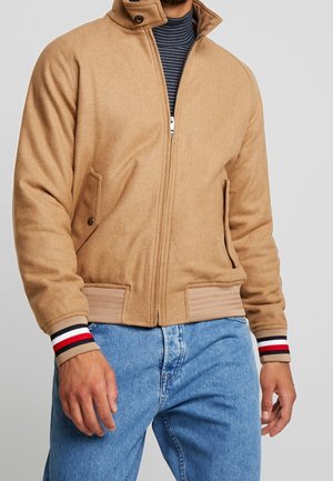 Man wearing a tan zip-up jacket with striped cuffs over a navy striped shirt and light blue jeans, standing against a gray background.