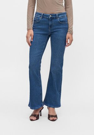 Femme portant un jean évasé bleu, un haut beige à manches longues et des sandales marron à talons ouverts, se tenant devant un fond blanc uni.