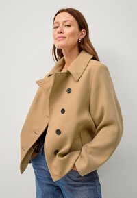 Tan uldblandingsjakke med bred krave, dobbeltræksdesign, fem sorte knapper og sidelommer; båret over blå denimjeans.