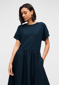 Φόρεμα σκούρου μπλε με δομημένο πάνω μέρος και ρέον, πιεσμένο κάτω μέρος. Βραχέα μανίκια, στρογγυλή λαιμόκοψη και πλευρικές τσέπες είναι ορατά.