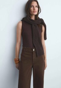 Top sin mangas marrón con una bufanda texturizada de color marrón oscuro. El conjunto incluye pantalones marrones de pierna ancha y alta cintura, además de una pulsera gruesa.