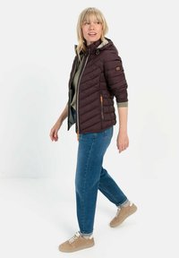 Padded bordeauxfarbige Jacke mit Kapuze, diagonal gesteppt, mit Reißverschlusstaschen und Lederakzenten, kombiniert mit blauen Jeans und beigen Schuhen.