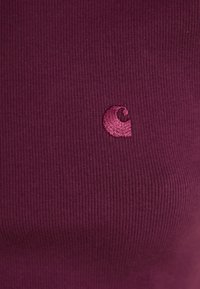 Primo piano di un tessuto a coste di un profondo viola con un piccolo logo ricamato in maroon nella zona del petto.