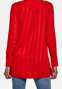 Camicia rossa a maniche lunghe con righe verticali, polsini con bottoni e orlo curvo. Tessuto leggero e semitrasparente con colletto e spalla.
