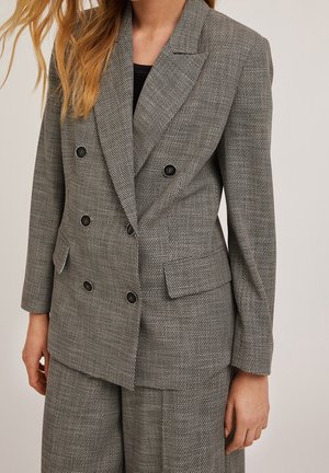 Femme portant un blazer gris texturé à double boutonnage et un pantalon assorti sur un haut noir, debout devant un fond uni.