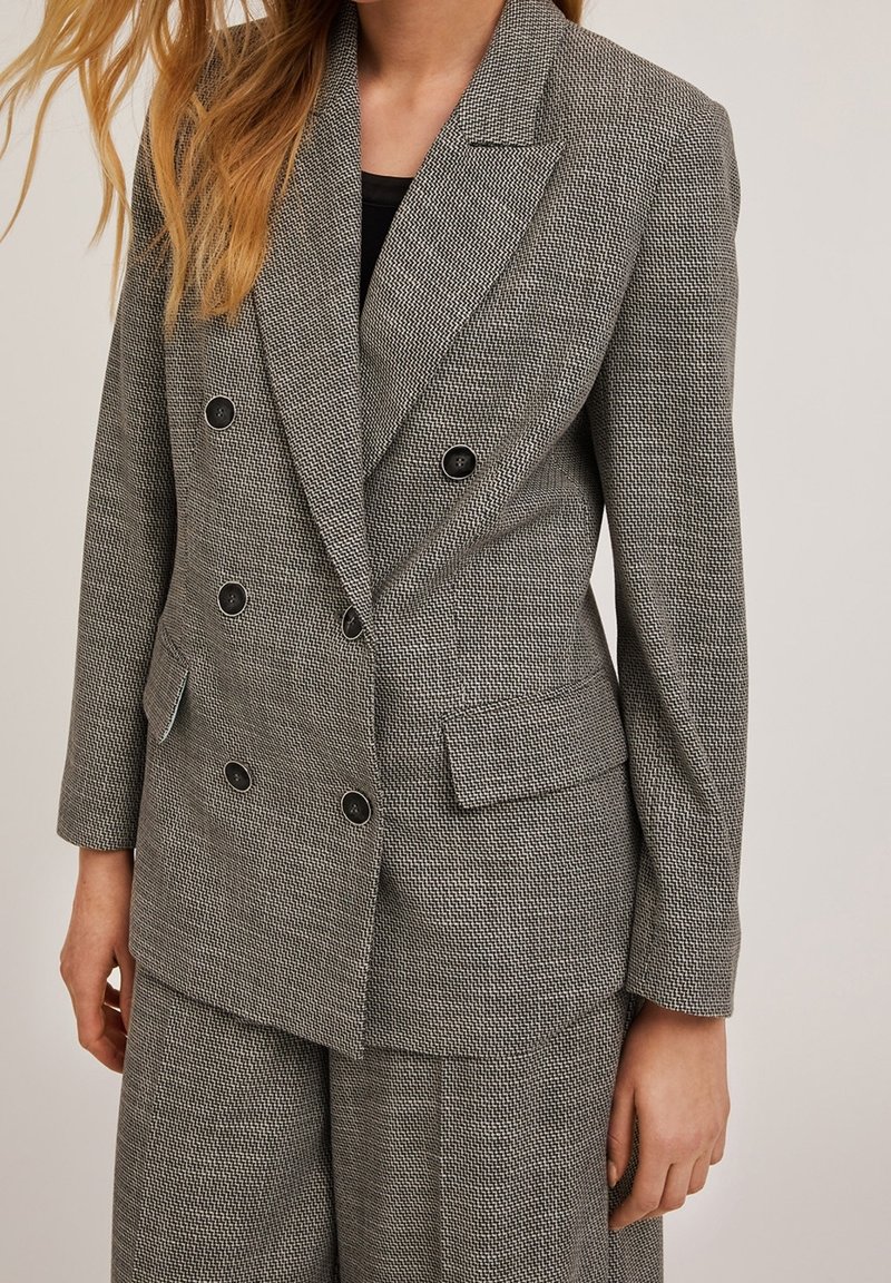 Femme portant un blazer gris texturé à double boutonnage et un pantalon assorti sur un haut noir, debout devant un fond uni.