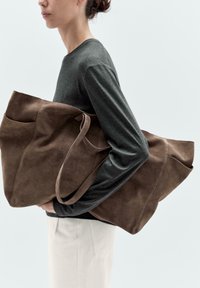 Brun mocka tote-väska med två handtag, en stor ytterficka och en mjuk, texturerad yta. Modellen håller den mot en neutral bakgrund.