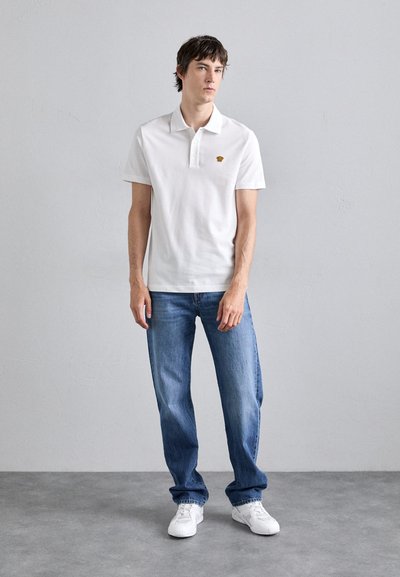 Chemise polo blanche à manches courtes et col, avec un petit logo jaune. Associée à un jean en denim bleu et des baskets blanches.