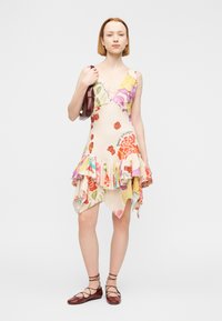 Φλοράλ φόρεμα από ελαφρύ ύφασμα με βολάν στο τελείωμα, διακοσμημένο με πολύχρωμα τριαντάφυλλα και λεπτομέρειες. Το μοντέλο κρατά μια μπορντό τσάντα και φορά καφέ δερμάτινα παπούτσια με κορδόνια.