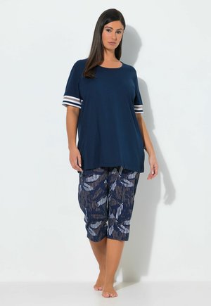 Femme aux longs cheveux foncés portant un t-shirt ample bleu marine à manches courtes rayées et un pantalon capri bleu marine à motifs, debout pieds nus sur un fond uni.