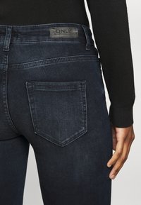 Jeans de ganga azul escuro com um bolso traseiro e uma etiqueta na cintura. Textura suave com um design ajustado, detalhe de costura mínimo.