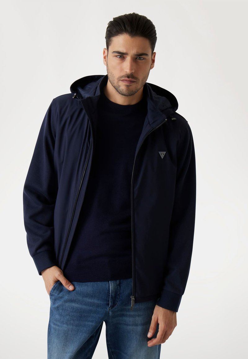 Giacca blu navy con zip frontale e cappuccio, caratterizzata da un logo discreto sul petto. Texture morbida con polsini a coste e design dritto.