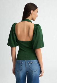 Top verde aderente con maniche a sbuffo e design con schiena scoperta. Il tessuto ha una leggera brillanza, abbinato a jeans blu con tasche posteriori.