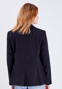 Blazer negro a medida con una única abertura trasera, hombros estructurados y puños abotonados. La tela tiene una textura suave y tejida.