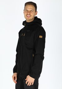 Black softshell jacket with a hood, featuring three zippered pockets and brown accents on the zippers and logo patch. Smooth texture.